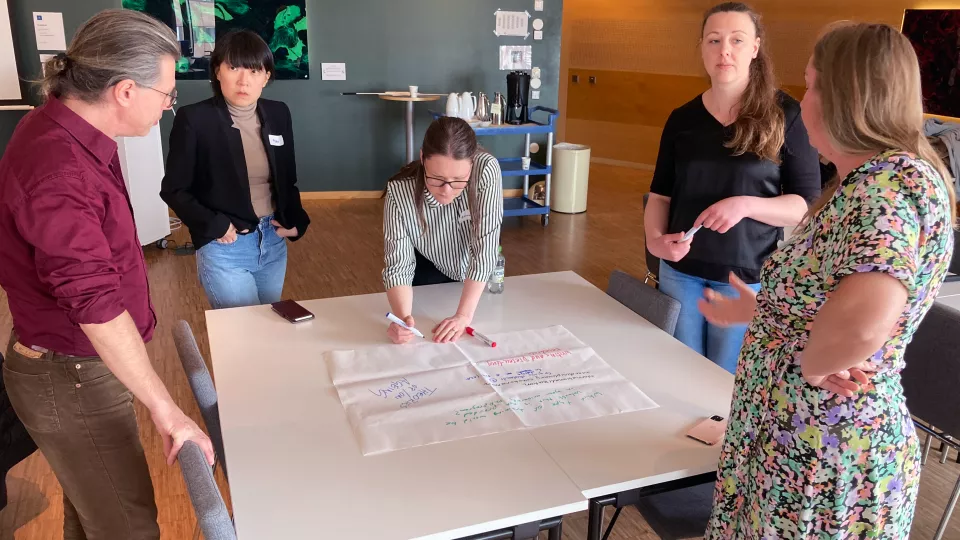 Fem personer står runt bord och diskuterar vad de ska skriva på pappret.